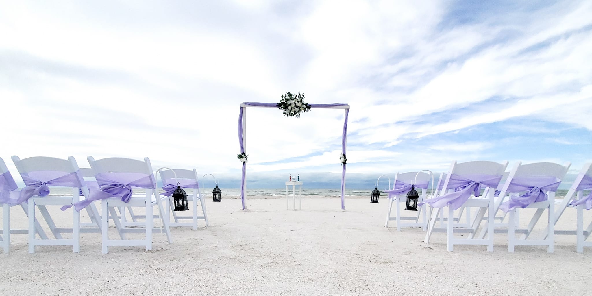 A beach wedding with white chairs and purple flowers.